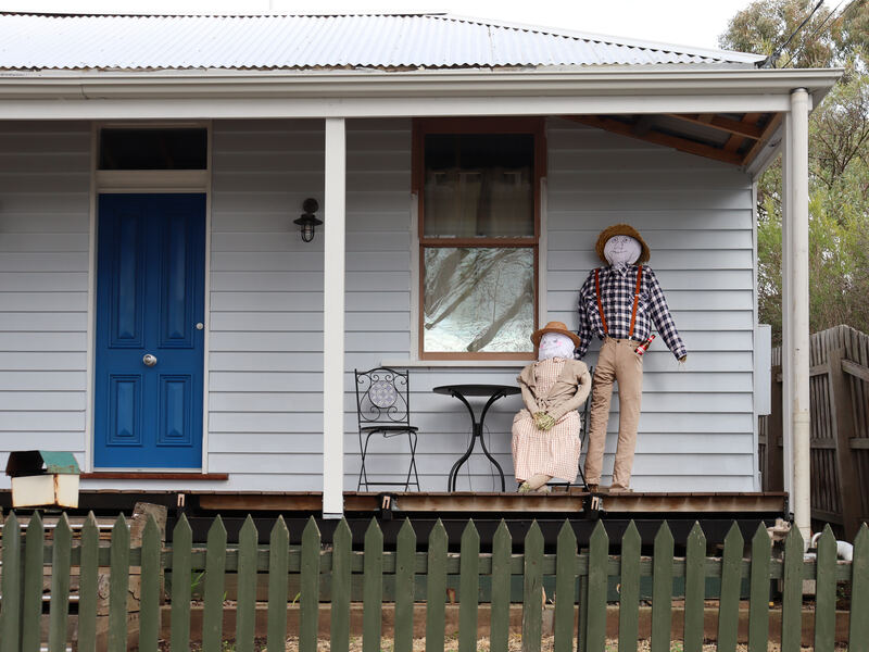 Two scarecrows sitting on the verandah of a weatherboard cottage